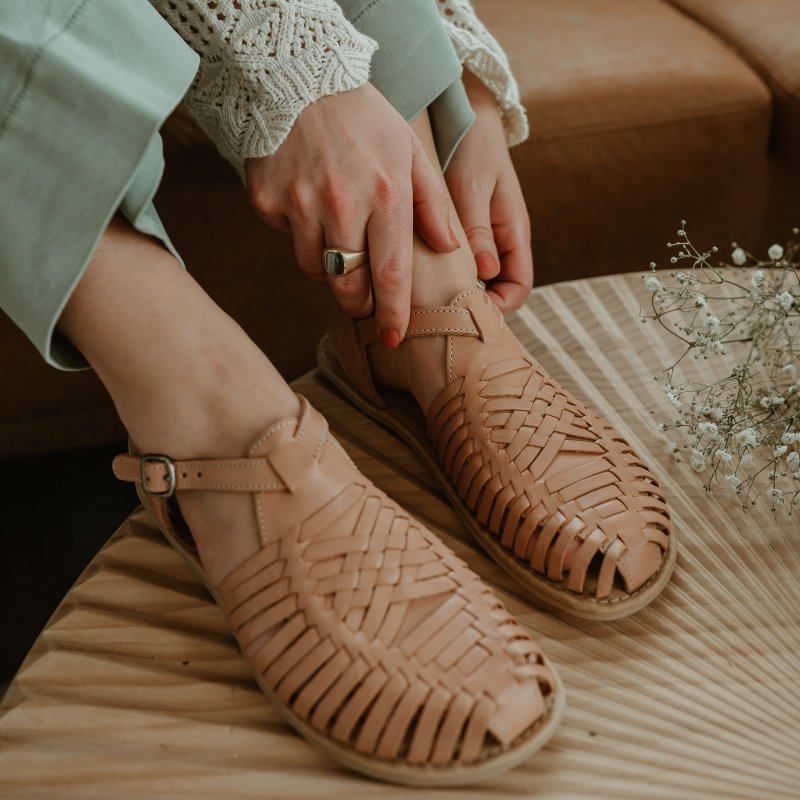 Close up of a woman's feet in Wide Huarache sandals in Tan leather adjustable the buckle with her feet propped up on a textured wood table.