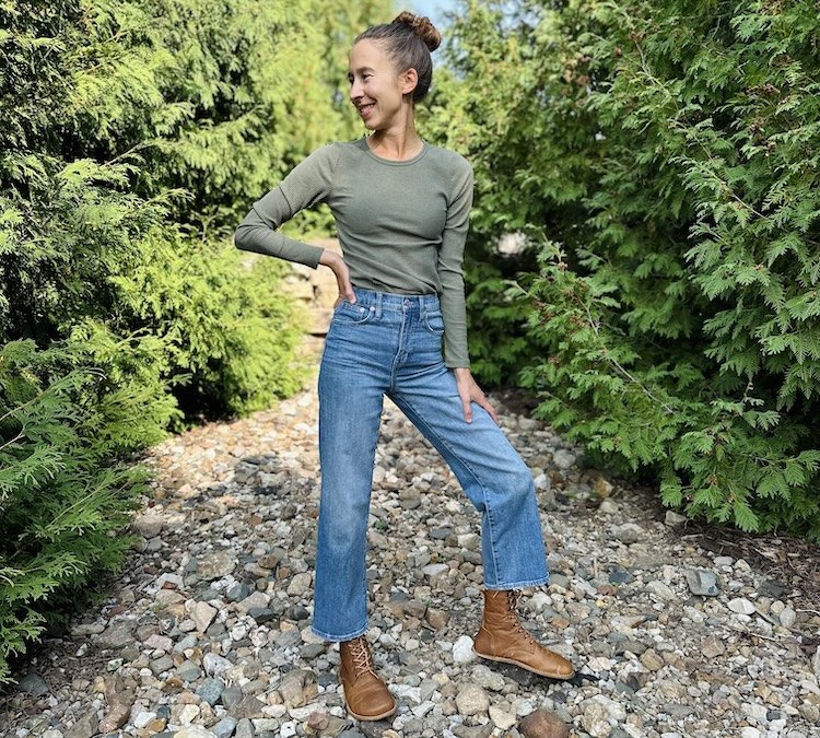 A woman standing outside in nature wearing denim and Origo Nomad Adventurer wide toe box barefoot boots in vintage brown, smiling and looking off in the distance