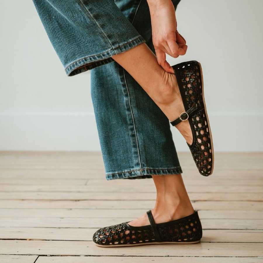 A close up knee down view of a woman's legs from the side. Her right foot is flat on the floor and her left is up in the air showing the zero drop outsole on her pair of black leather woven ballerina mary janes from Origo Shoes.