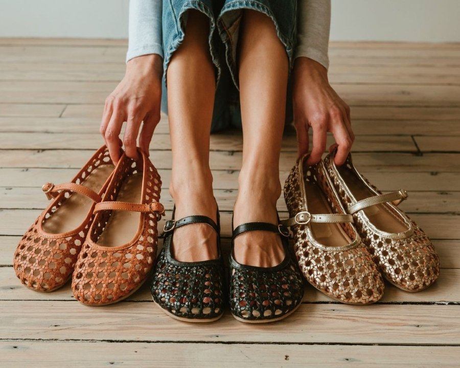 A woman holding 2 colors of Origo Shoes Woven Ballerina Mary Janes in her fingertips next to the pair of Black Woven barefoot ballerinas she has on her feet. The wide toe box is at the forefront of the photo.