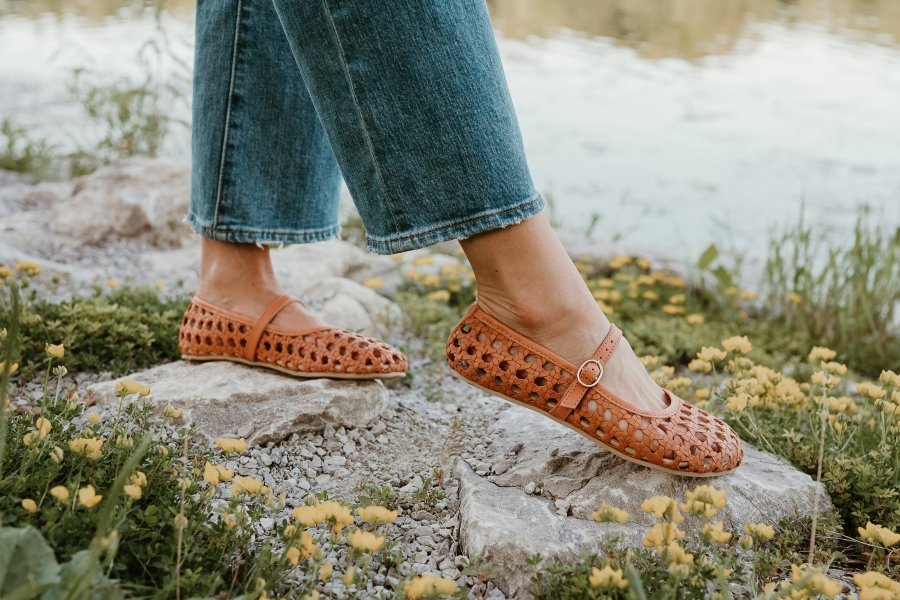 A side view of a pair of legs in wide jeans walking on rocks with yellow flowers around, wearing woven brown leather barefoot ballerinas from Origo Shoes