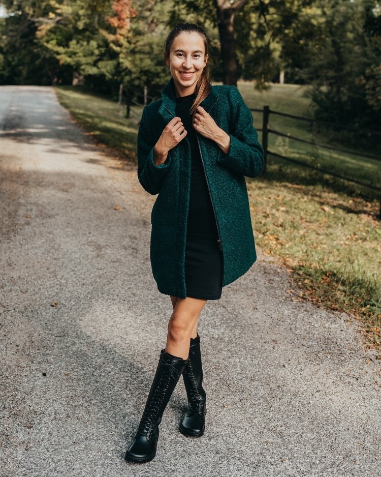 Full body view of a woman smiling at the camera wearing short black dress, and dark green overcoat being held at the lapel. She is wearing tall black lace up leather boots from Peerko called the Empire. They are sleek, stylish, and foot friendly.