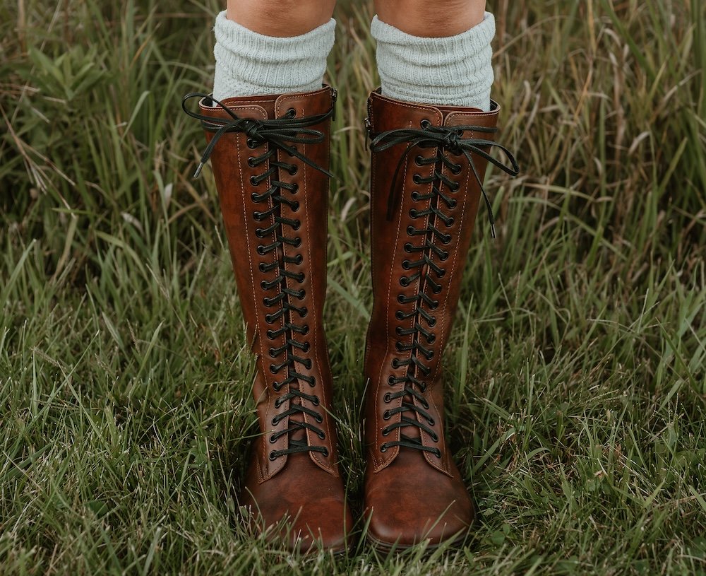 A close up from the front of a woman's lower legs standing in front of tall grass wearing grey boot socks and brown leather tall lace up barefoot boots - the Peerko Empire in brandy color.