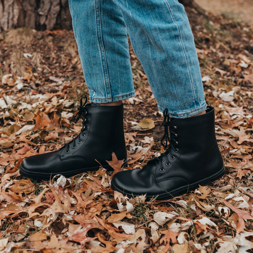 A pair of legs from the knee down walking outside on fall leaves wearing black lace up barefoot warm winter boots, the Peerko Frost. The feet are pointing to the left and the profile of the zero drop sole is facing the camera.