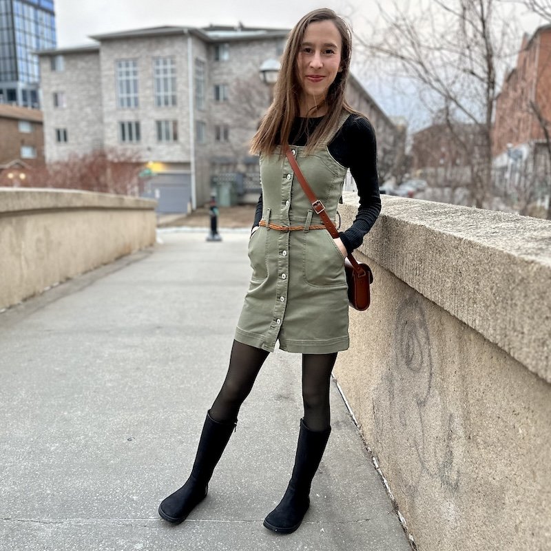 A woman standing outside smiling at the camera wearing a green dress, black stirrup tights, and black leather Peerko Regina tall barefoot boots