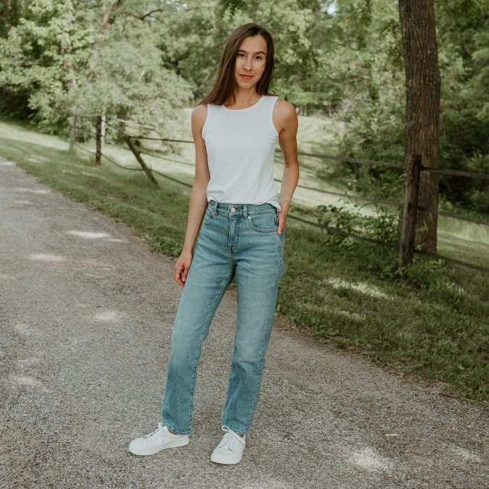 A woman standing on a road in jeans and a white tank, with her hand in her back pocket. She has Peerko white leather sneakers on her feet for comfortable all day wear.