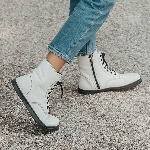 A close up of a pair of feet standing on pavement mid-walk wearing white leather Peerko Go wide toe box barefoot combat style lace up boots