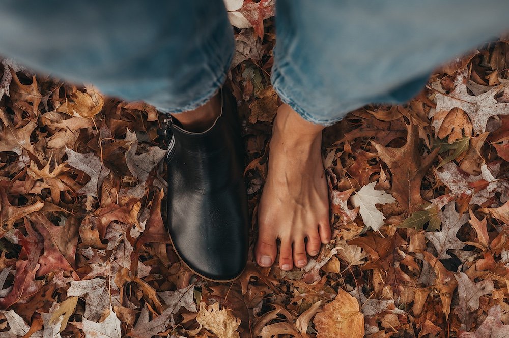 A top down view of a pair of feet standing in fallen autumn leaves. On one foot is a black barefoot ankle boot called the Rosa from Anya's Shop, and the other is a bare foot, to show that the boot mirrors the shape of the natural foot