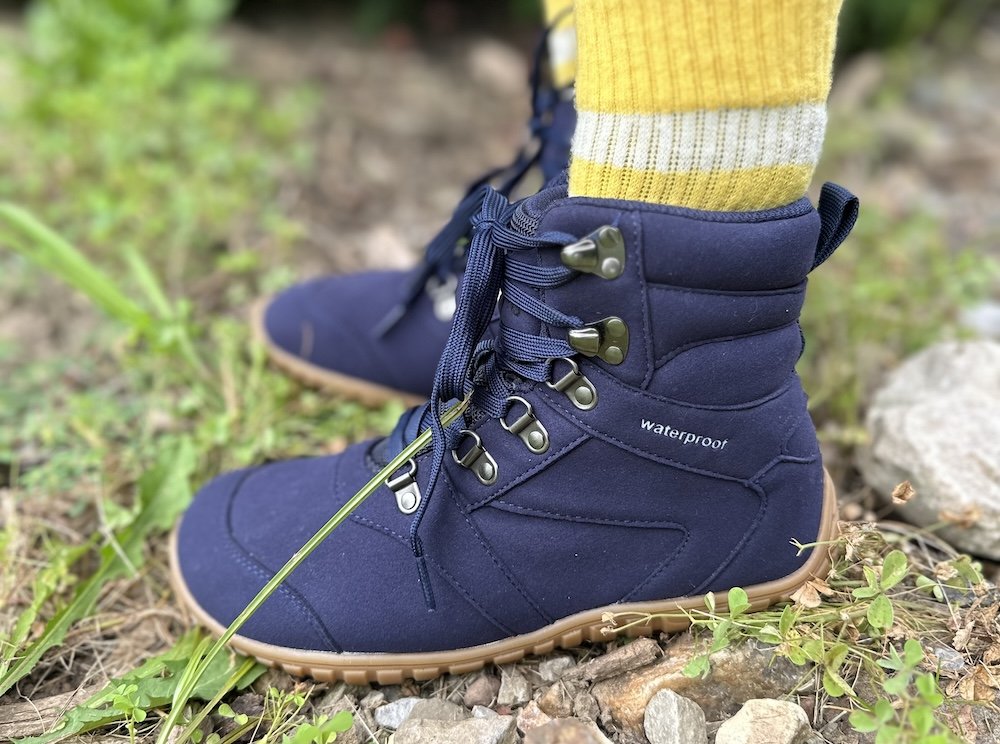 A close up side view of a person's feet standing outdoors on rocks and plants wearing Saguaro barefoot hiking boots in blue