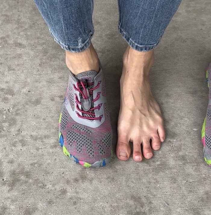 A top down view of a pair of feet standing on concrete. One is bare and the other is wearing Saguaro cheap barefoot sneakers