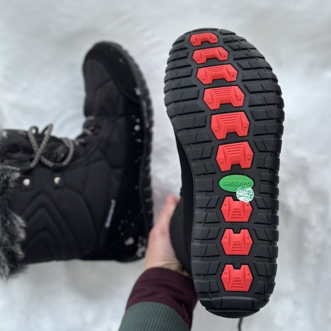 A hand holding a Saguaro Rise affordable barefoot snowboot to show the outsole. The other boot is in the background on it's side.