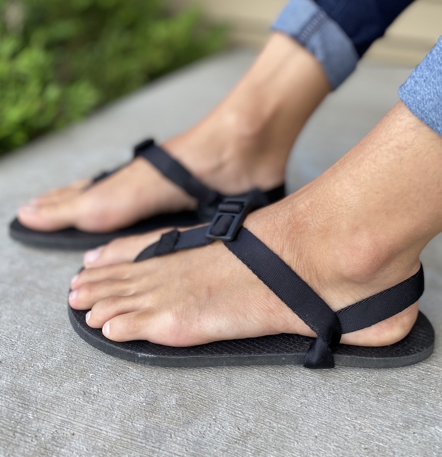 A side view of a pair of feet standing on concrete wearing black vegan Shamma Cruzers cheap barefoot sandals