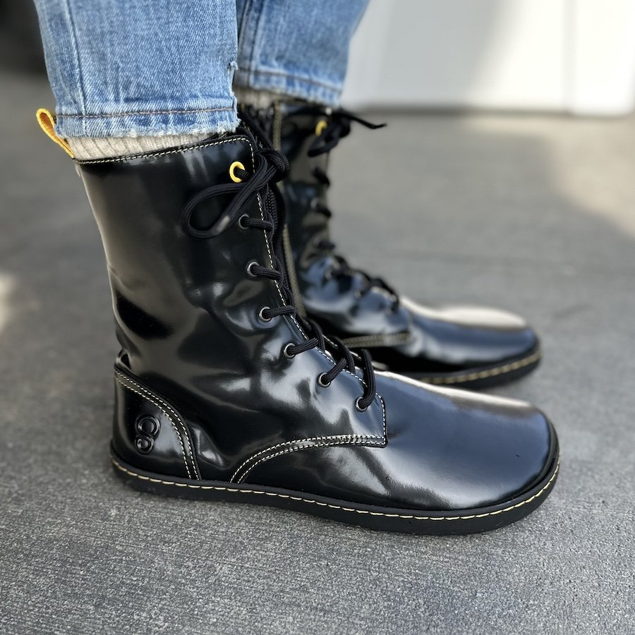 A close up side view of a pair of feet standing on concrete wearing new Shapen Barefoot Maverick boots in black shiny leather that look like Doc Martens