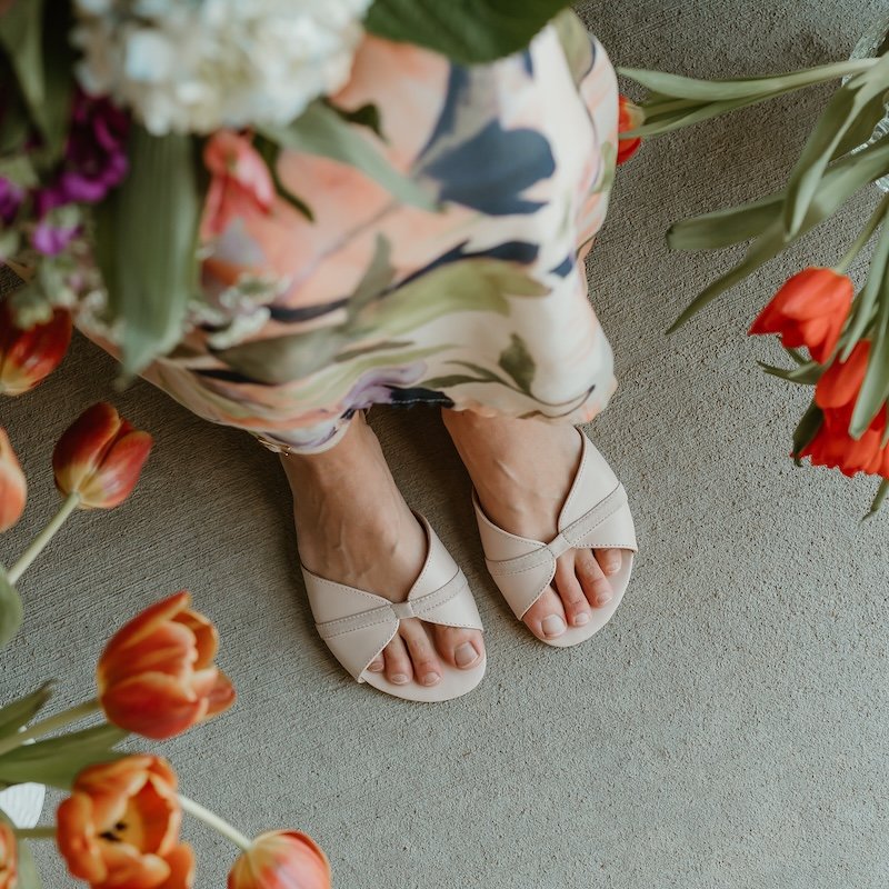 A pair of feet in Shapen Petal, barefoot dressy flats with a wide toe box. The woman is seated wearing a floral skirt, with tulips peeking into the frame at the sides