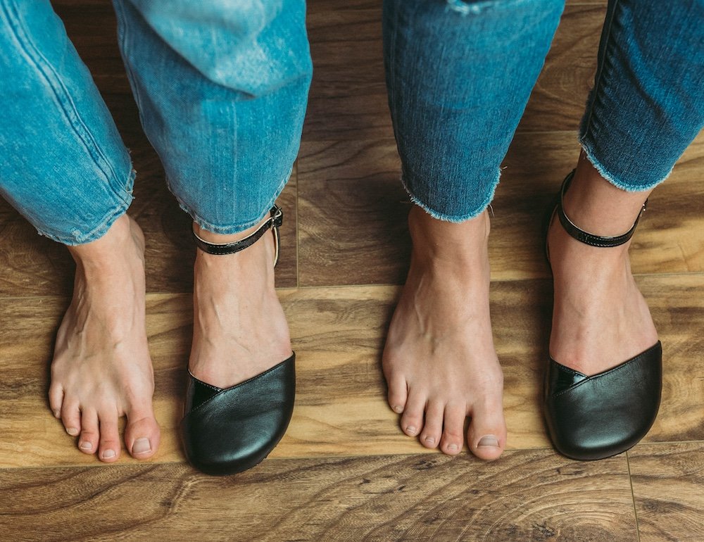 a top down view of two women's feet standing on a wood floor. The left pair of feet have one foot bare and one wearing a black Shapen Poppy barefoot women's dress shoe. The right pair of feet has one foot bare and the other wearing a SHapen Poppy barefoot women's dress shoe in black, wide width to show how the different widths fit different foot types.