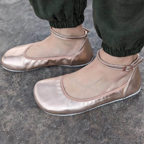 Side profile of a pair of feet wearing metallic Shapen Tulip 3 barefoot comfortable dress flats for extra wide feet with a matching ankle strap and anatomical toe box. The foot in back is slightly forward and dark green pants are visible at the ankles.