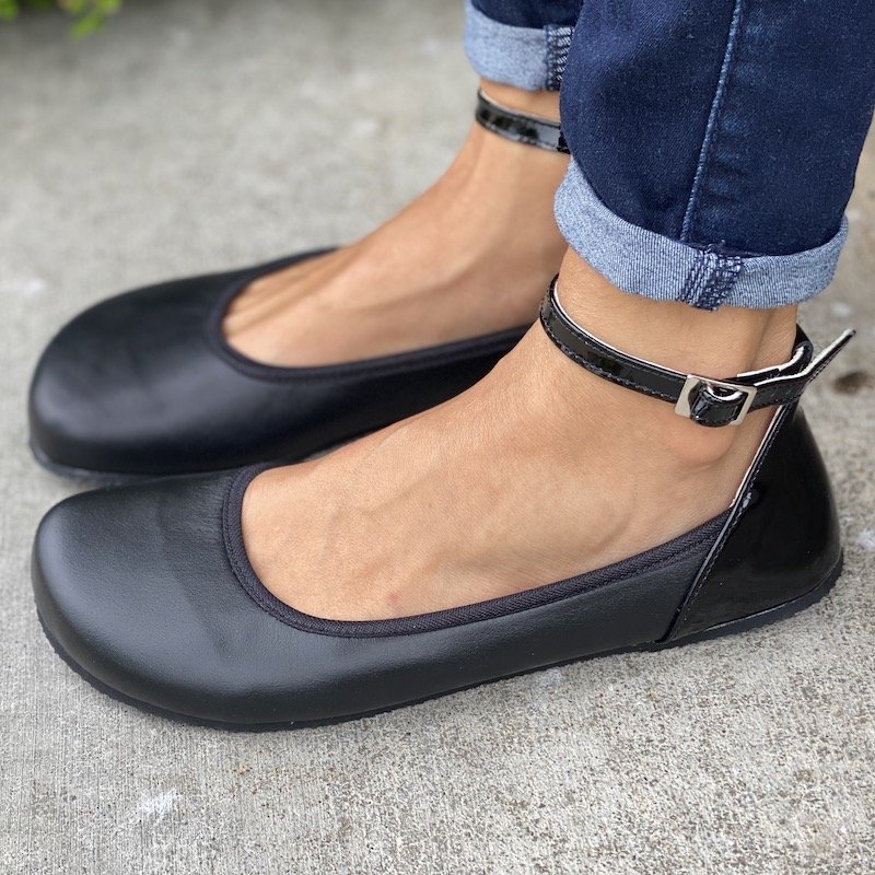 a side view of the black Shapen barefoot tulip flats worn by a woman standing on concrete.