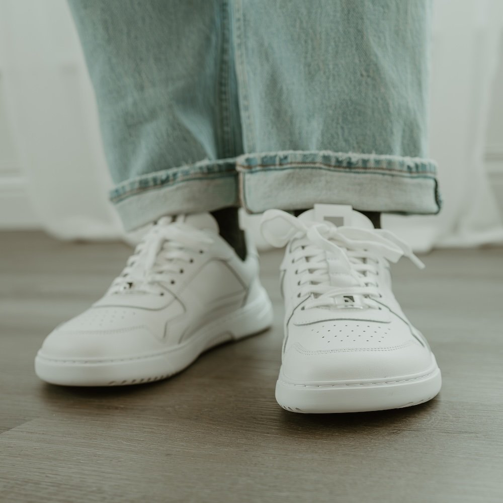 A front view of a person's legs in cuffed light colored denim, and a pair of white Barebarics leather Zing 2 sneakers with a wide toe box and low stack height flat sole.
