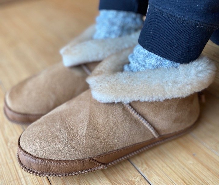 A side view of a pair of feet standing on wood floor wearing Softstar Shoes wool sheepskin Fireside slippers in Wide width with cozy socks peeking out the top