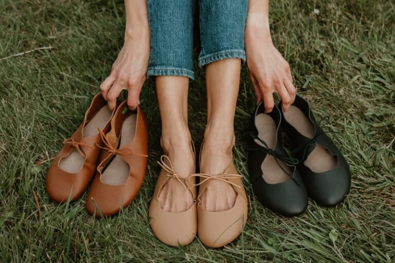 A knee down view of a woman seated on the grass wearing macadamia colored Softstar Vintage Tie Ballerine flats. She is holding the other 2 collection colors - Nutmeg and Black.