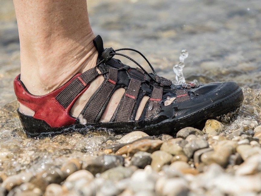 A close up of a single foot stepping on the rocky shore of a body of water wearing Sole Runner FX Training barefoot sandals in red and brown