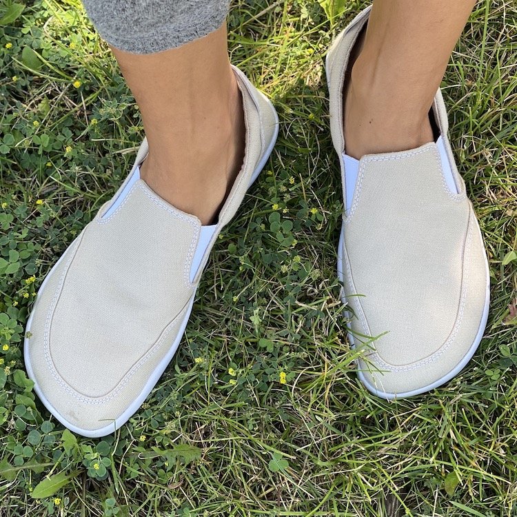 A pair of feet standing on grass wearing Splay Shoes Freestyle SLip on barefoot sneakers in tan canvas