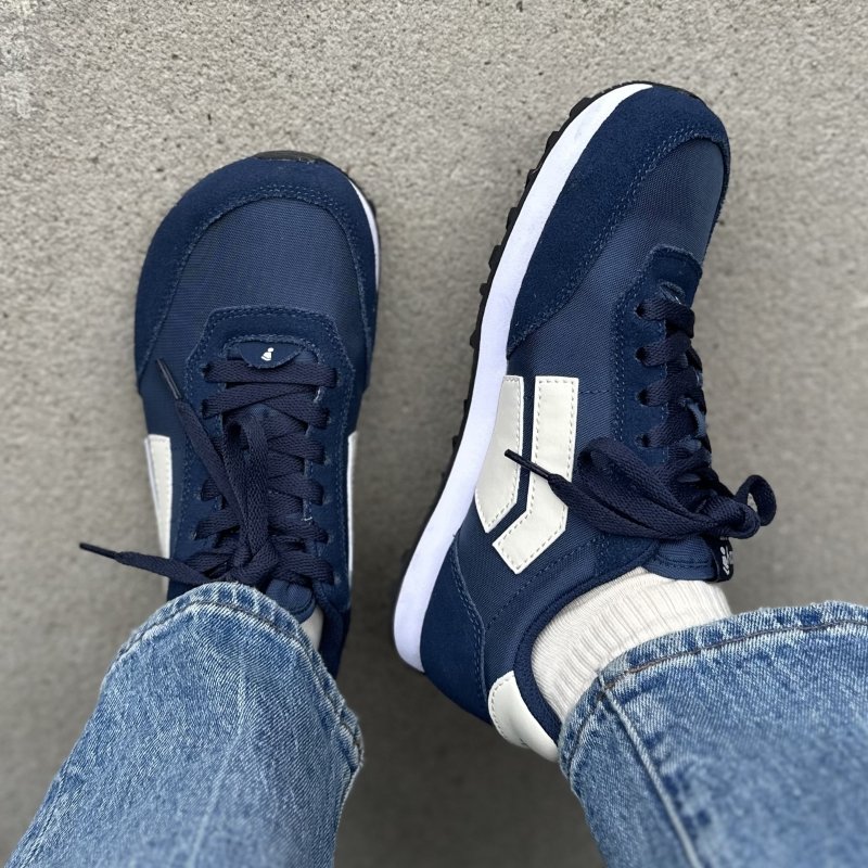 Splay Runner sneakers with a retro vibe in dark blue with cream accents. The right foot is angling the shoe out to the side to show the zero drop outsole, and the left is flat on the ground highlighting the wide toe box.