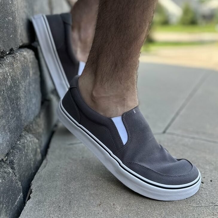 Side close up view of a man's feet wearing Splay Streetstyle slip on barefoot sneakers that look like Vans. The back foot is propped up against a stone wall and the forefoot is flexed.