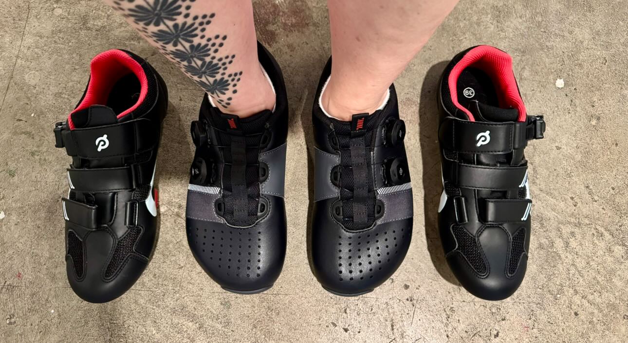 A person's feet in anatomical cycling shoes from Strong Feet Athletics standing with conventional narrow pointy cycling shoes to the sides to show the shape comparison