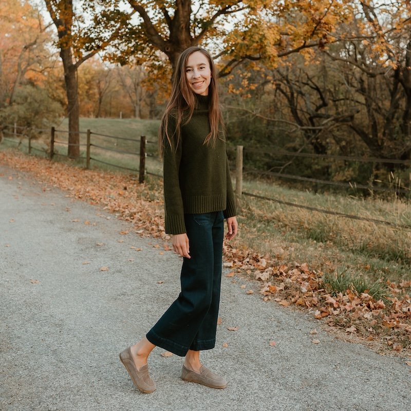 A woman standing out in the street among fall colored leaves wearing a sweater, jeans, and wide toe box Dalia barefoot loafers from Anya's Shop in taupe nubuck color.