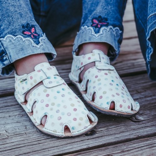 A young girl in embroidered denim wearing white Tikki Aranya barefoot leather sandals for kids with colorful polka dots