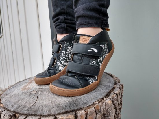 A close up of a kids feet wearing Tikki Moon black leather sneakers in Graffiti print standing on a log on their tiptoes showing the easy flexibility of these super cool barefoot shoes for kids