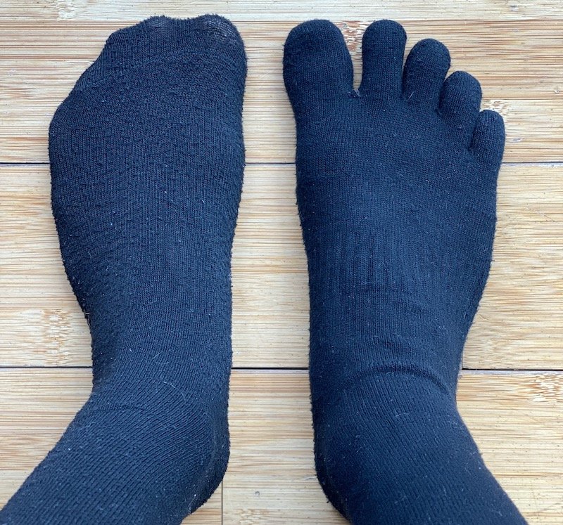 A top down view of a pair of feet standing on wood floor - the left foot is wearing a traditional black sock with the toes squished together. The right foot is wearing a black toe sock with the toes splayed out