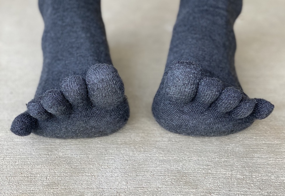 A close up of a pair of feet standing on concrete wearing dark grey toe socks with the toes lifted up and splayed
