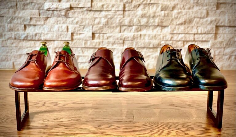 A shoe rack in front of a stone wall containing 3 pairs of shiny wide toe box men's zero drop dress shoes. All 3 pairs of Tomas Sullivent dress shoes are lined up facing the camera in light brown, dark brown, and black.