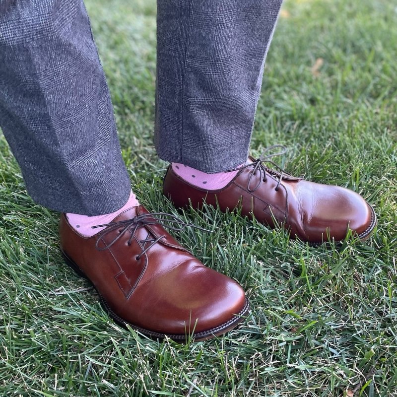 A shin down side view of a man in subtle plaid dress pants, pink polka dot socks, and Tomas Sullivent wide toe box dress shoes in rich brown leather