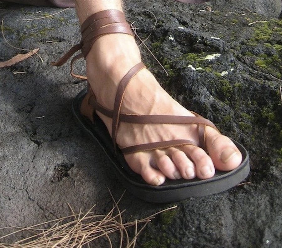 A strappy brown leather sandal. Straps are thin and wrap around the ankle and are for men or women. Shown on a mans foot atop a rock.