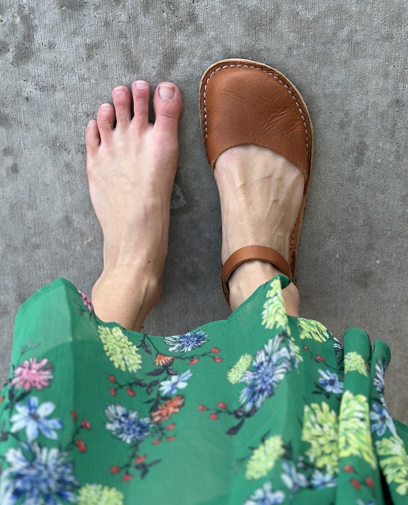 A close up of a pair of feet standing on concrete - the left foot is bare and the right foot is wearing Crupon Sandals Trevi barefoot in Ambra with a natural foot shape. the hem of a green floral skirt is visible.