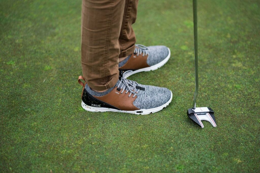A side view of a person standing on a golf green wearing True Linkswear OG Feel barefoot golf shoes with a golf club next to his feet