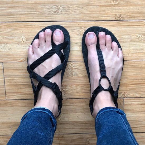 a top down view of a pair of feet with one foot in an Unshoes Uinta sandals (black) and the other in an Unshoes Keota Sandal (black).