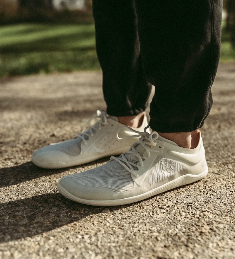 A close up of a man's legs and feet standing on pavement wearing white Vivobarefoot Primus Lite barefoot sneakers