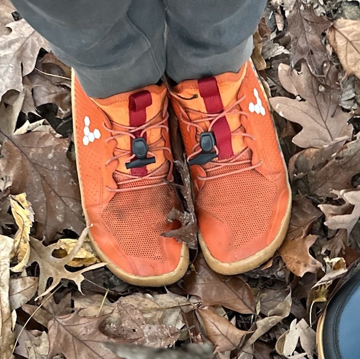 A pair of vibrant orange Vivobarefoot mesh sneakers for kids standing on dried leaves