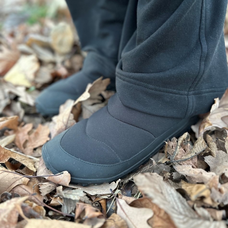 A close up front angled view of a person in black pants wearing Vivobarefoot explorer Mid Barefoot winter boots on dead leaves.