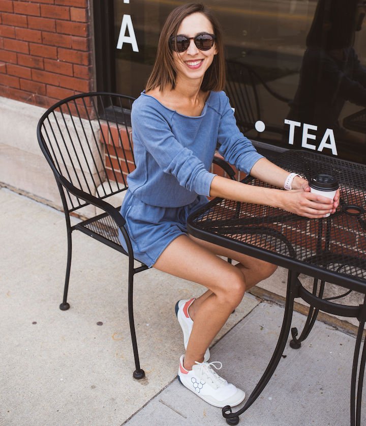 A woman at a cafe table holding a coffee, wearing a blue romper, and Vivobarefoot Geo Court sneakers.