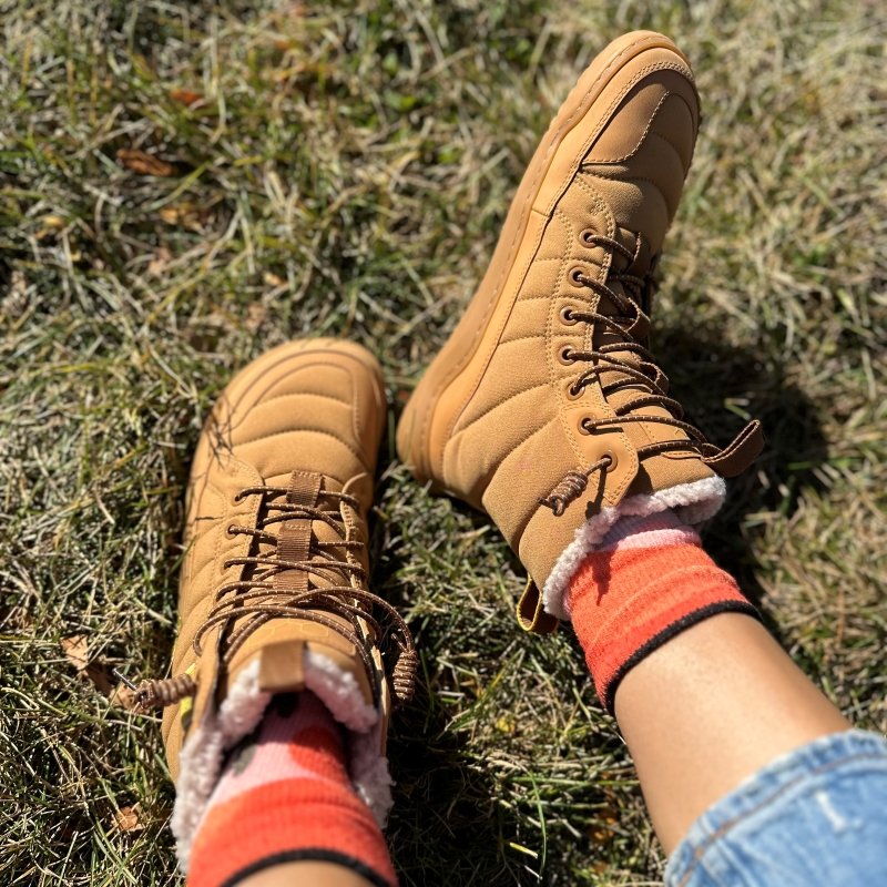 A pair of legs relaxing outside wearing Vivobarefoot Gobi Hiber puffer barefoot boots with elastic laces, a wide toe box, and flexible flat outsole