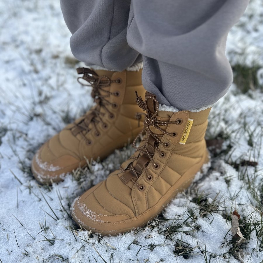 A pair of Vivobarefoot Gobi Hiber puffer winter insulated warm barefoot boots in camel standing on snowy ground.