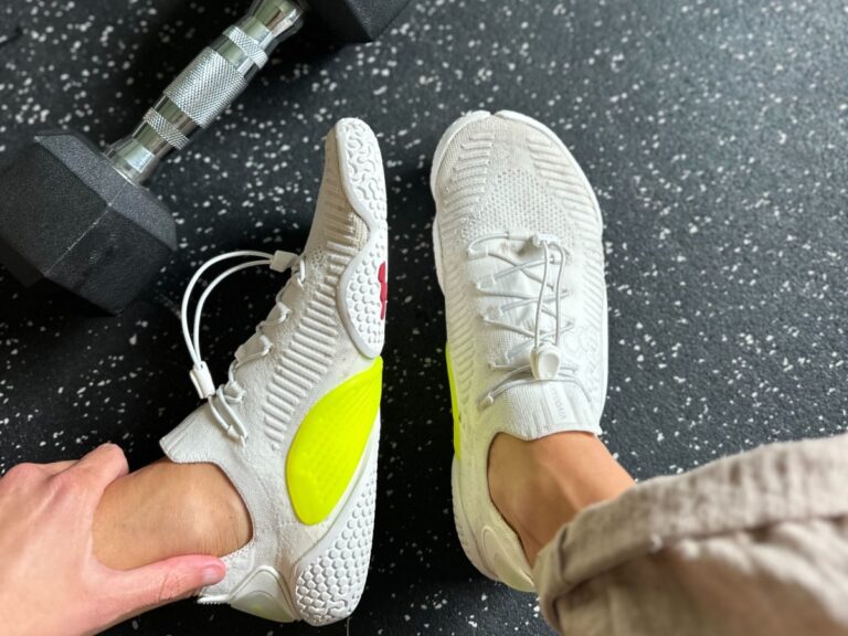 Close up top down and side view of a person's legs wearing Vivobarefoot Motus Flex on a gym floor with a weight in the corner of the frame