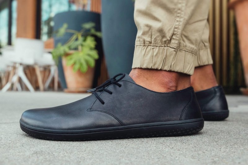 Side close up view of a man in khaki chino jogger pants with Vivobarefoot Ra men's barefoot dress shoes in black high quality leather on a zero drop outsole