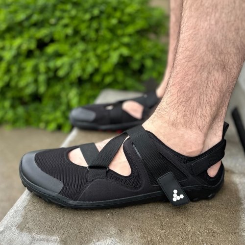Side close up view of black closed toe barefoot Vivobarefoot Tracker sandals with a wide toe box. The man wearing the sandals in standing on wet concrete and the Z strap is visible on the top of the foot