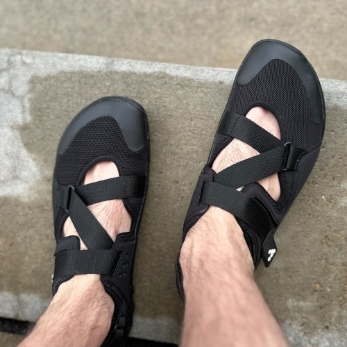 Top view of Vivobarefoot Tracker Sandals being worn by a man standing on wet concrete steps. The reinforced wide toe box and Z style strap are the focus of the photo.